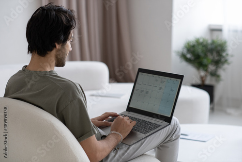 Young contemporary man with laptop on knees looking through working plan on screen while sitting on sofa in living room
