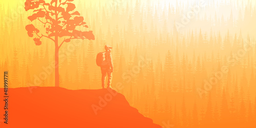 A tourist with a backpack on a rock meets dawn. Mountains covered by forest, taiga. Morning haze, the light of sunrise.