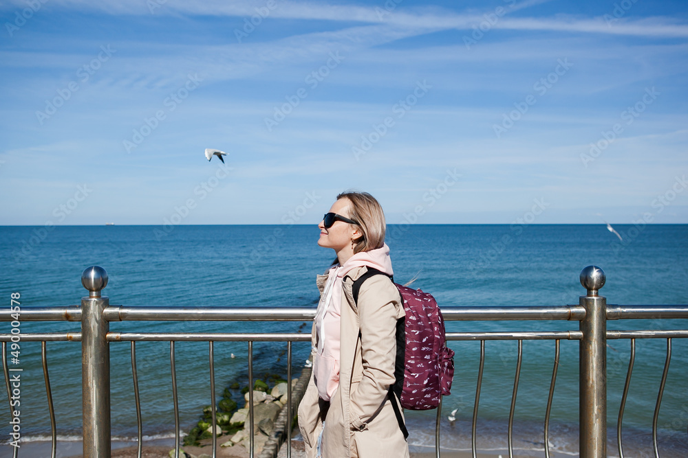 a blonde girl with a backpack on the background of a cold sea, seagulls in the sky. the concept of travel and trips