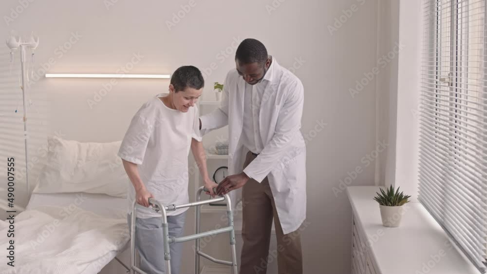 Medium shot of African American male doctor helping mature Caucasian woman to stand up from hospital bed using walkers, looking at window in hospital ward and talking about post surgery recovery Medium shot of African American male doctor helping mature Caucasian woman to stand up from hospital bed using walkers, looking at window in hospital ward and talking about post surgery recovery