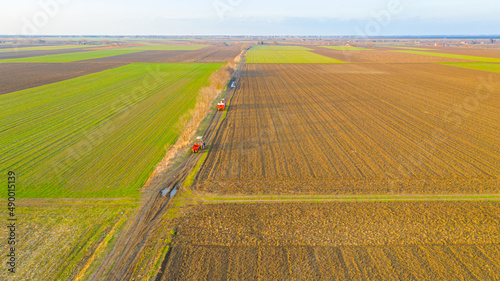 Aerial view of two tractors, one with mounted trailer and other has agricultural machine for spreading artificial fertilizer as travel on dirty road