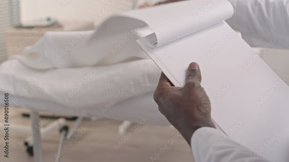 Slowmo closeup of unrecognizable African American doctor filling in medical documents of female patient lying in oxygen mask in hospital bed in blurred background Slowmo closeup of unrecognizable African American doctor filling in medical documents of female patient lying in oxygen mask in hospital bed in blurred background