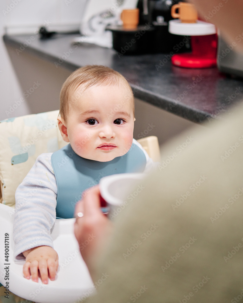 Mother giving a yogurt to her son sitting in the highchair Mother giving a yogurt to her son sitting in the highchair