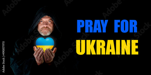 PRAY FOR UKRAINE.  Monk in a hood with a heart of Ukraine in his hands on a black background