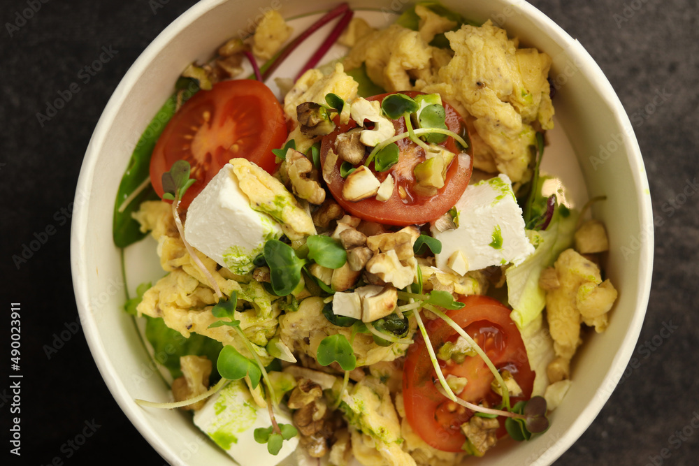 Bowl with scramble, feta cheese, tomatoes, walnuts and microgreens on a dark background in an eco paper box. Healthy food delivery concept.