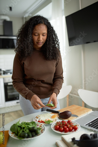 Beautiful casually dressed dark-skinned girl with curly fuzzy black hair cooking breakfast  decorating plate with fried eggs with spinach leafs  avocado and cherry tomatoes  standing against kitchen