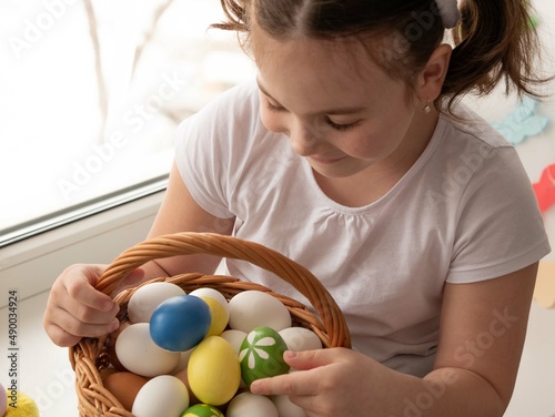 A little girl in a white sweater and two ponytails is sitting on the windowsill happy holding a basket full of colored Easter eggs. Easter concept photo