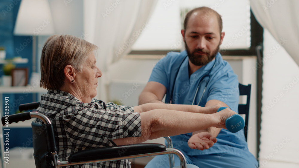 Nurse assisting disabled patient with physical exercises for recovery. Retired woman sitting in wheelchair using dumbbells for healing activity in nursing home while nurse giving support Nurse assisting disabled patient with physical exercises for recovery. Retired woman sitting in wheelchair using dumbbells for healing activity in nursing home while nurse giving support