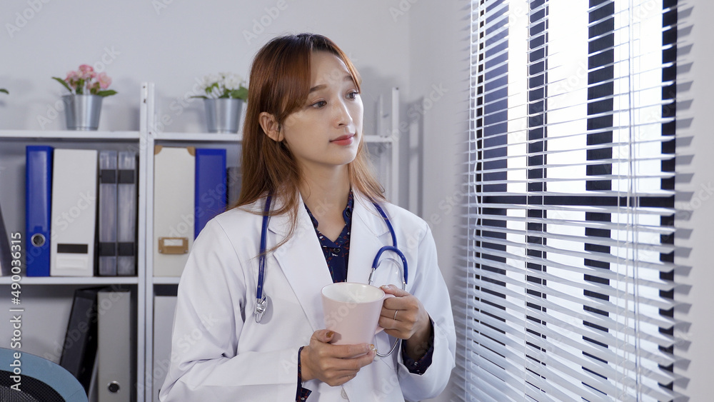 Taiwanese female in medical uniform is looking into distance with a smile while drinking tea by office window and enjoying the tranquil morning. Taiwanese female in medical uniform is looking into distance with a smile while drinking tea by office window and enjoying the tranquil morning.