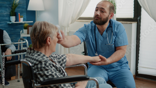 Medical assistant using thermometer on old disabled woman in nursing home. Retired patient in wheelchair while nurse measuring temperature and fever for healthcare recovery checkup