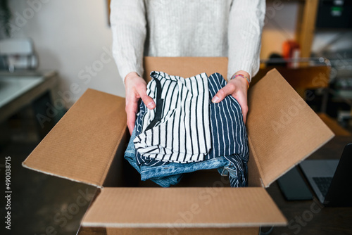 Business Woman prepares a package in a cardboard box for shipping with clothes from her online store - Millennial sells second-hand used clothing in her home - Start up concept
