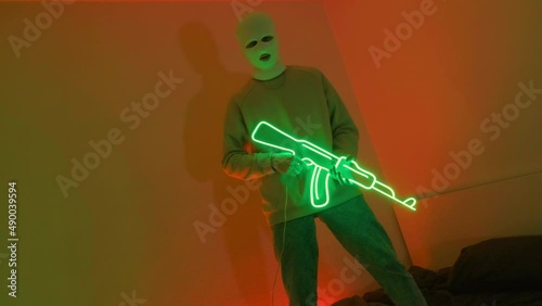 A gangster in a white mask holds a neon machine gun. A young guy is standing in an ultraviolet room on a bed. Video 4k for a rap clip in a neon room.