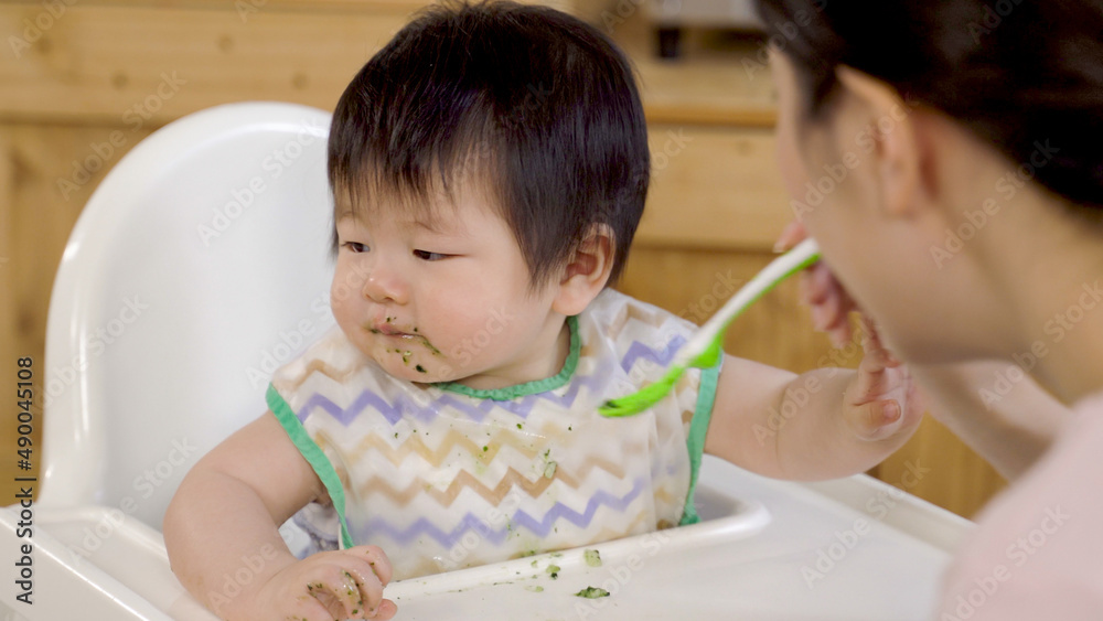 innocent asian young toddler sitting in the baby chair at home is looking away while being fed with pureed food by her mom at breakfast. innocent asian young toddler sitting in the baby chair at home is looking away while being fed with pureed food by her mom at breakfast.