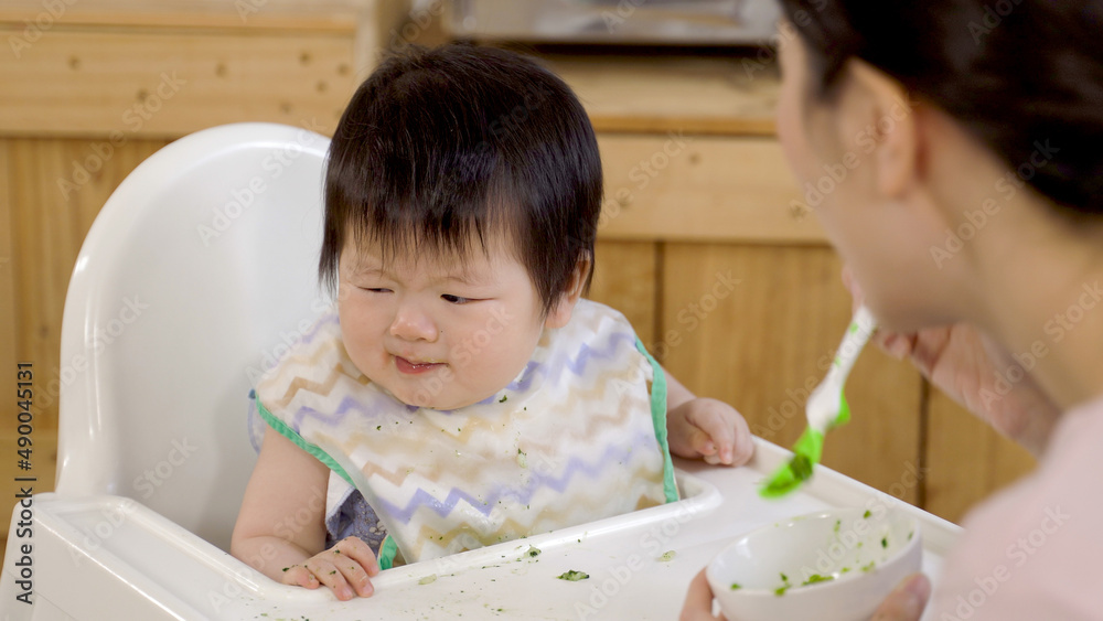close up irritated and upset asian baby is resisting her mother from wiping the food away from her mouth with a napkin on chair after meal at home. close up irritated and upset asian baby is resisting her mother from wiping the food away from her mouth with a napkin on chair after meal at home.
