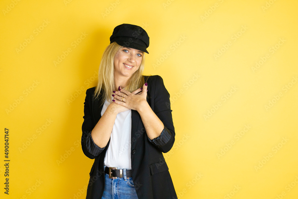 Beautiful woman wearing casual white t-shirt and a cap over isolated yellow background smiling with her hands on her chest and grateful gesture on her face. Beautiful woman wearing casual white t-shirt and a cap over isolated yellow background smiling with her hands on her chest and grateful gesture on her face.