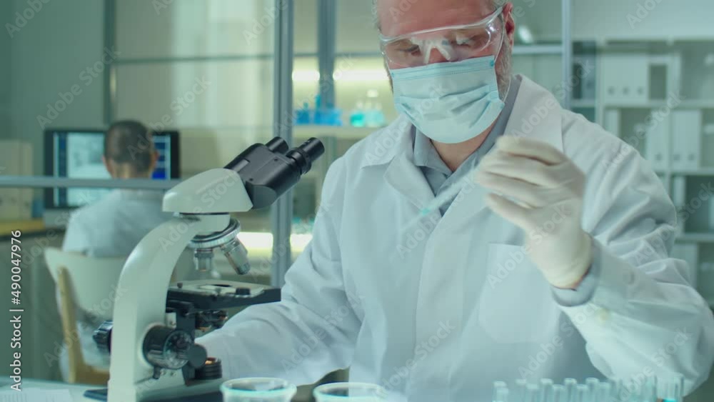 Senior Caucasian chemist in protective mask, gloves and glasses putting specimen sample on glass slide and examining it under microscope while working in laboratory Senior Caucasian chemist in protective mask, gloves and glasses putting specimen sample on glass slide and examining it under microscope while working in laboratory