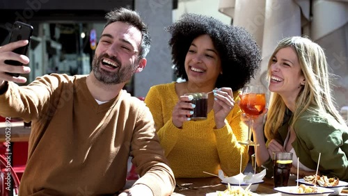 Video of bearded man taking selfie with female friends while having drink at terrace restaurant. photo