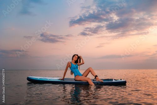 A young smiling tanned woman poses sitting on a sup board. Sunset in the background. Copy space. Concept of sup boarding