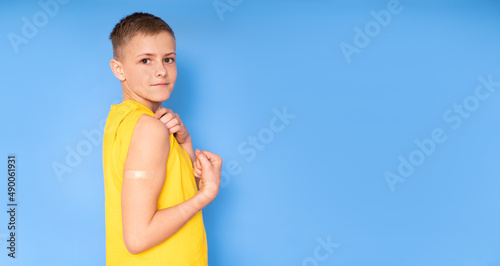 A teenage boy with a patch on his arm clenches his fist and bends his biceps after vaccination. Injectable vaccine against covid, healthcare for children. place for text