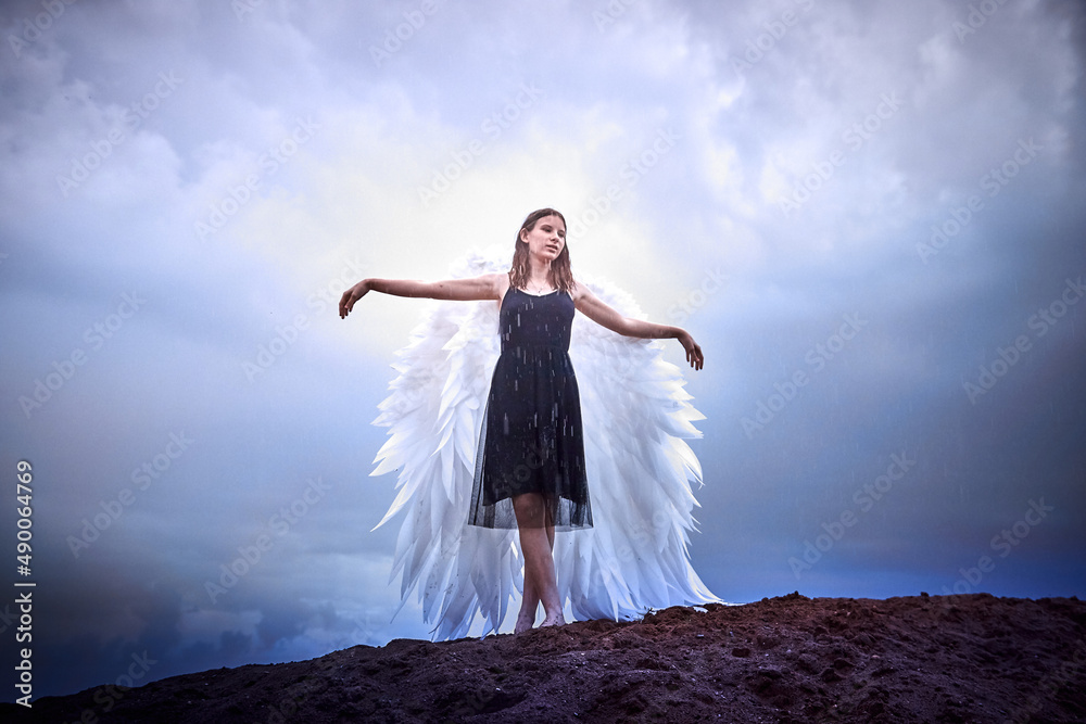 Young slim girl in black dress with with white wings dances on sand dunes against a dramatic sky before a thunderstorm. Model posing during photo shoot on nature. Beautiful angel in rain day Young slim girl in black dress with with white wings dances on sand dunes against a dramatic sky before a thunderstorm. Model posing during photo shoot on nature. Beautiful angel in rain day