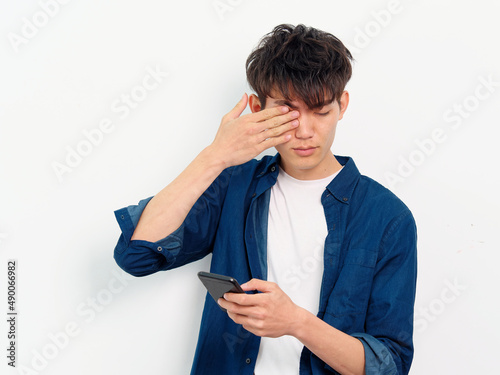 Portrait of handsome Chinese young man with black curly hair in blue shirt posing against white wall background. Fingers rubbing eyes with mobile phone in hand, looks tired, front view studio shot. photo