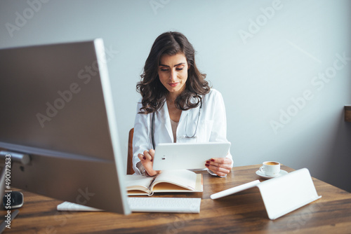 Portrait of adult female doctor sitting at desk in office clinic. One happy Young Woman or Doctor Nurse in Hospital Clinic Office using tablet PC. Female doctor working on her tablet pc photo