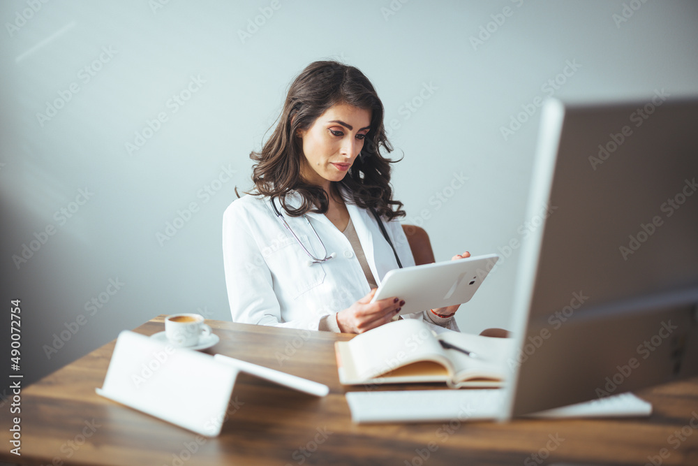 Portrait of adult female doctor sitting at desk in office clinic. One happy Young Woman or Doctor Nurse in Hospital Clinic Office using tablet PC. Female doctor working on her tablet pc Portrait of adult female doctor sitting at desk in office clinic. One happy Young Woman or Doctor Nurse in Hospital Clinic Office using tablet PC. Female doctor working on her tablet pc
