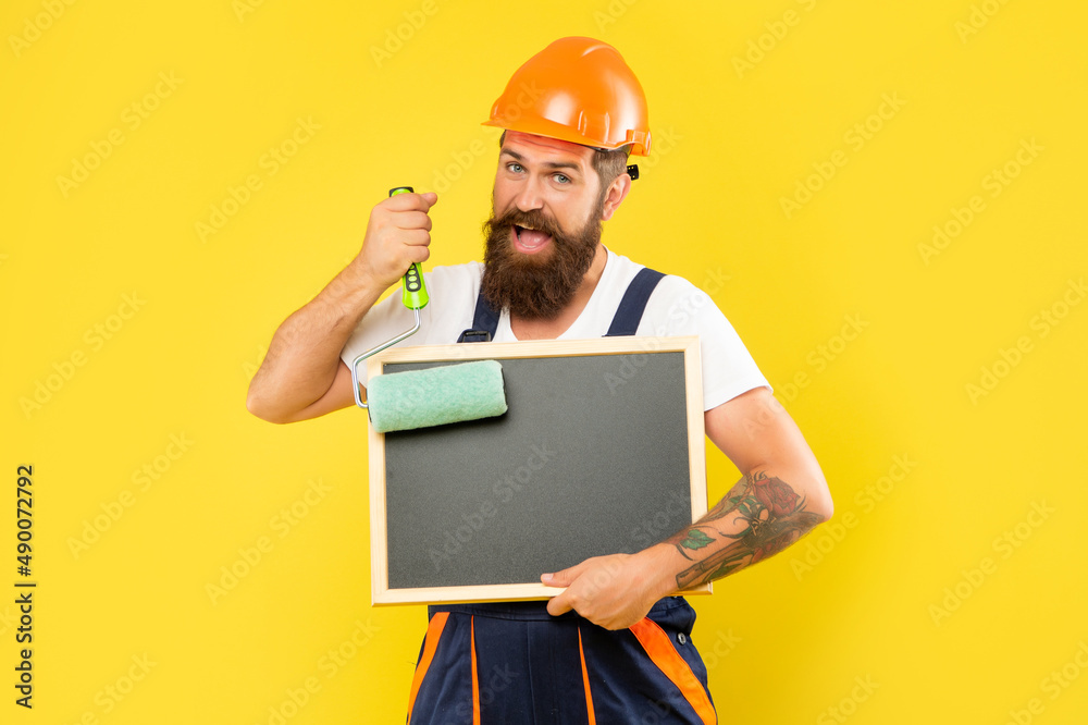 smiling man housepainter in helmet and work clothes hold paint roller and blackboard smiling man housepainter in helmet and work clothes hold paint roller and blackboard