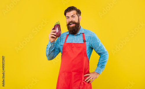 Happy man in red apron holding eggplant yellow background, grocer photo