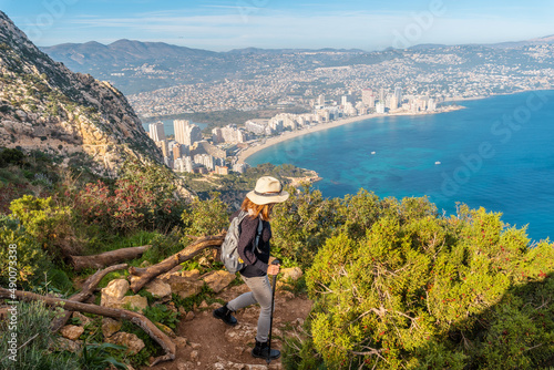 A young hiker wearing a hat on the descent path of the Penon de Ifach Natural Park with the city of Calpe in the background, Valencia. Spain. Mediterranean sea. View of La Fossa beach