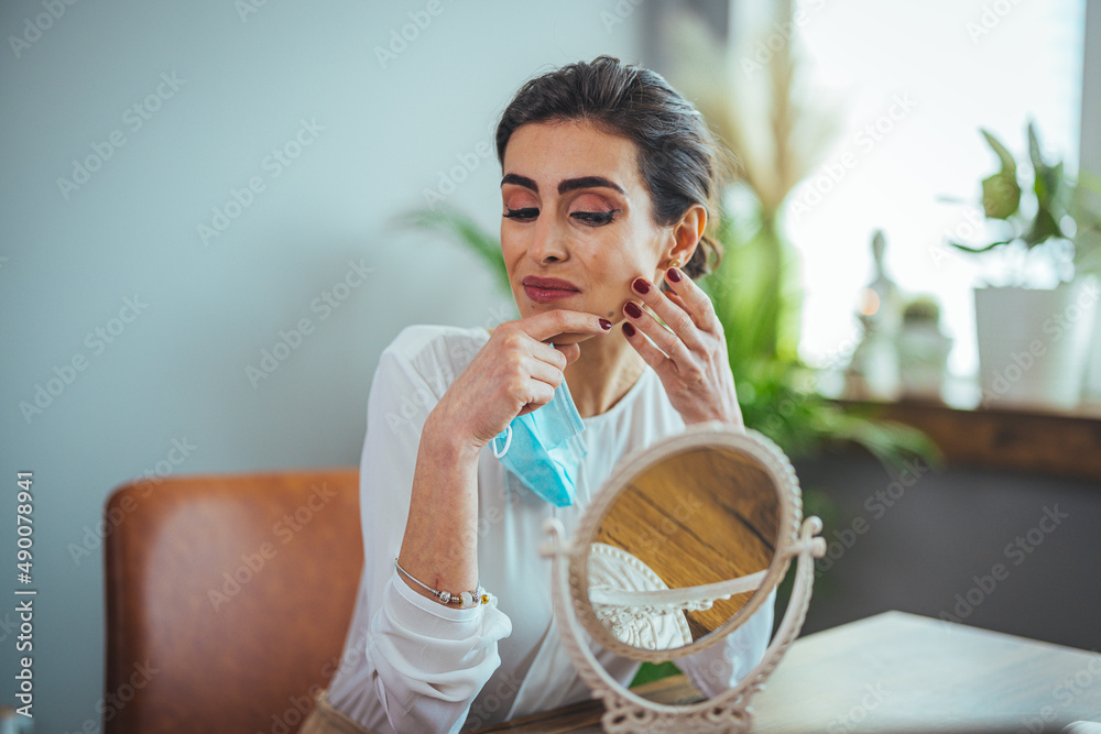 Young beautiful woman looking herself in the mirror at home. Worried about acne caused by wearing a mask. Maskne. Problems with acne. Young beautiful woman looking herself in the mirror at home. Worried about acne caused by wearing a mask. Maskne. Problems with acne.