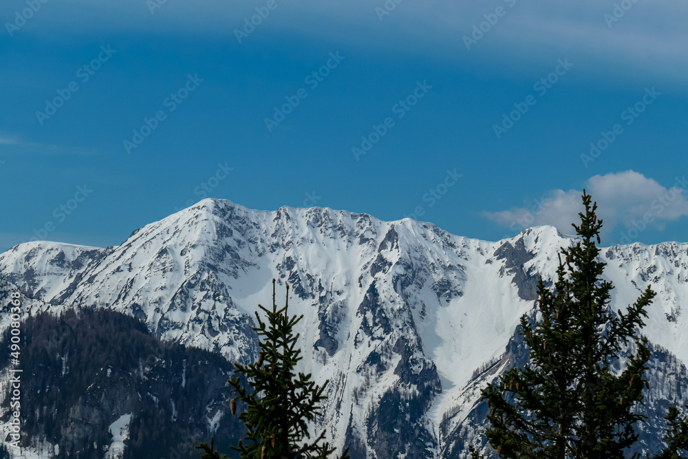 Scenic view of snow capped mountain peaks of Karawanks on the way to Sinacher Gupf in Carinthia, Austria. Mount Wertatscha is visible through dense forest in early spring. Rosental on a sunny day.Hike