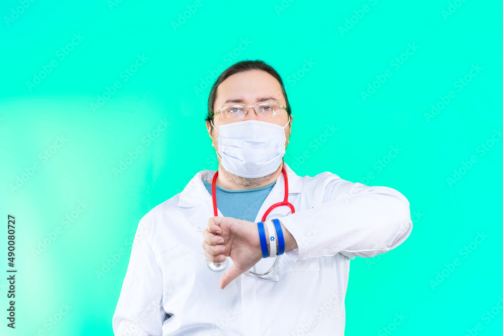 A doctor in a white coat and mask points his thumb down. The doctor shows a gesture of disapproval. Bracelet in the colors of the flag of Israel. A doctor in a white coat and mask points his thumb down. The doctor shows a gesture of disapproval. Bracelet in the colors of the flag of Israel.