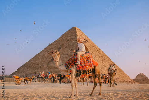 Happy Tourist man with hat riding on camel background pyramid of Egyptian Giza, sun light Cairo, Egypt
