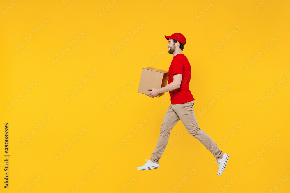 Full body side profile view delivery guy employee man in red cap T-shirt uniform workwear work as dealer courier jump high hold cardboard box run fast jump high isolated on plain yellow background Full body side profile view delivery guy employee man in red cap T-shirt uniform workwear work as dealer courier jump high hold cardboard box run fast jump high isolated on plain yellow background