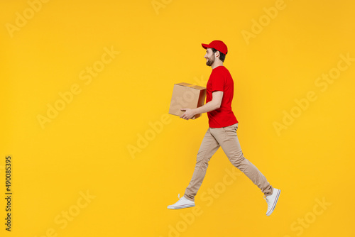 Full body side profile view delivery guy employee man in red cap T-shirt uniform workwear work as dealer courier jump high hold cardboard box run fast jump high isolated on plain yellow background