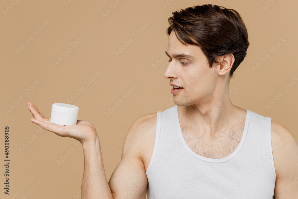 Caucasian young man 20s perfect skin in undershirt hold moisturizer facial cream container isolated on pastel pastel beige background studio portrait. Skin care healthcare cosmetic procedures concept.
