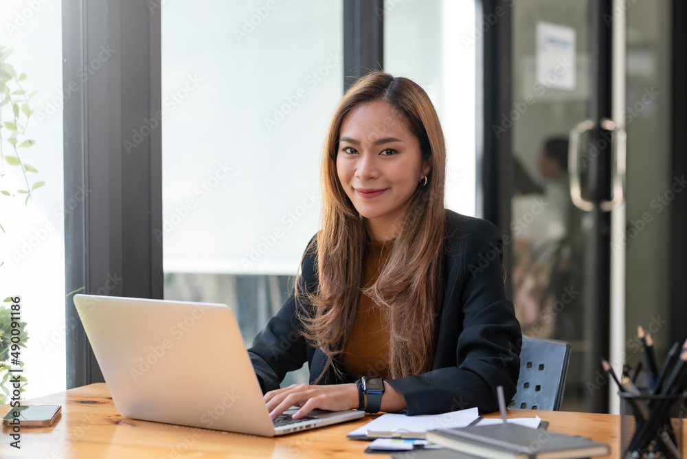 Portrait of smiling young beautiful Asian businesswoman sitting with laptop computer looking at camera in the office.