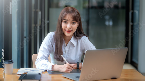 Portrait of smiling young beautiful Asian businesswoman sitting with laptop computer looking at camera in the office.