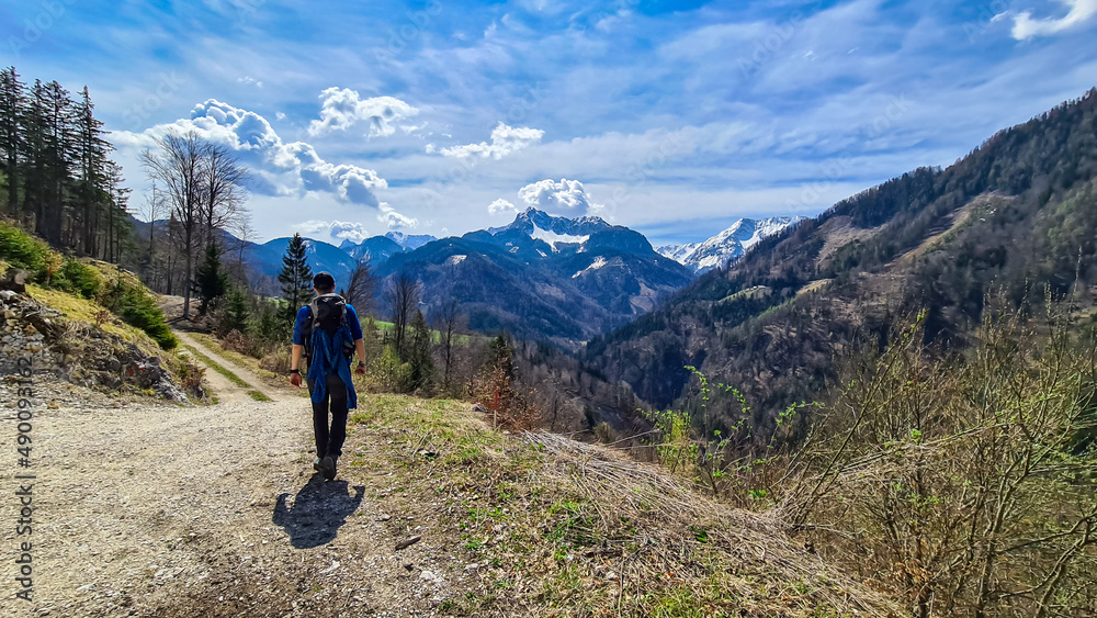 Man with backpack and scenic view of snow capped mountain peaks of Karawanks near Sinacher Gupf in Carinthia, Austria. Mount Hochstuhl (Stol) visible through forest in spring. Rosental sunny day. Awe
