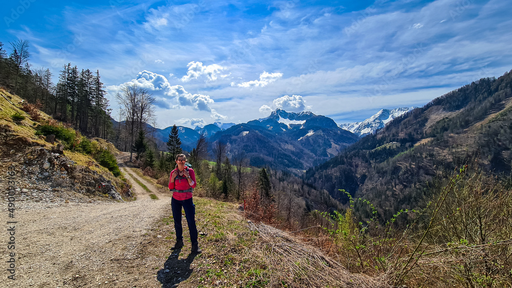 Woman with backpack and scenic view of snow capped mountain peaks of Karawanks near Sinacher Gupf in Carinthia, Austria. Mount Hochstuhl (Stol) visible through forest in spring. Rosental sunny day.Awe