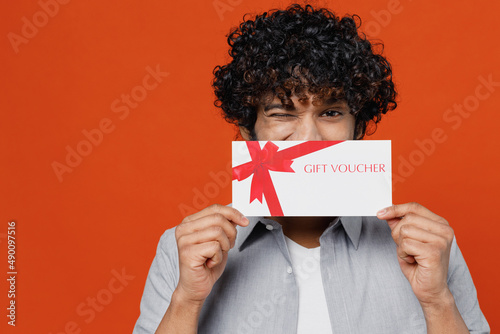 Young bearded Indian man 20s years old wears blue shirt hold cover close hiding mouth with gift certificate coupon voucher card for store blinking isolated on plain orange background studio portrait.