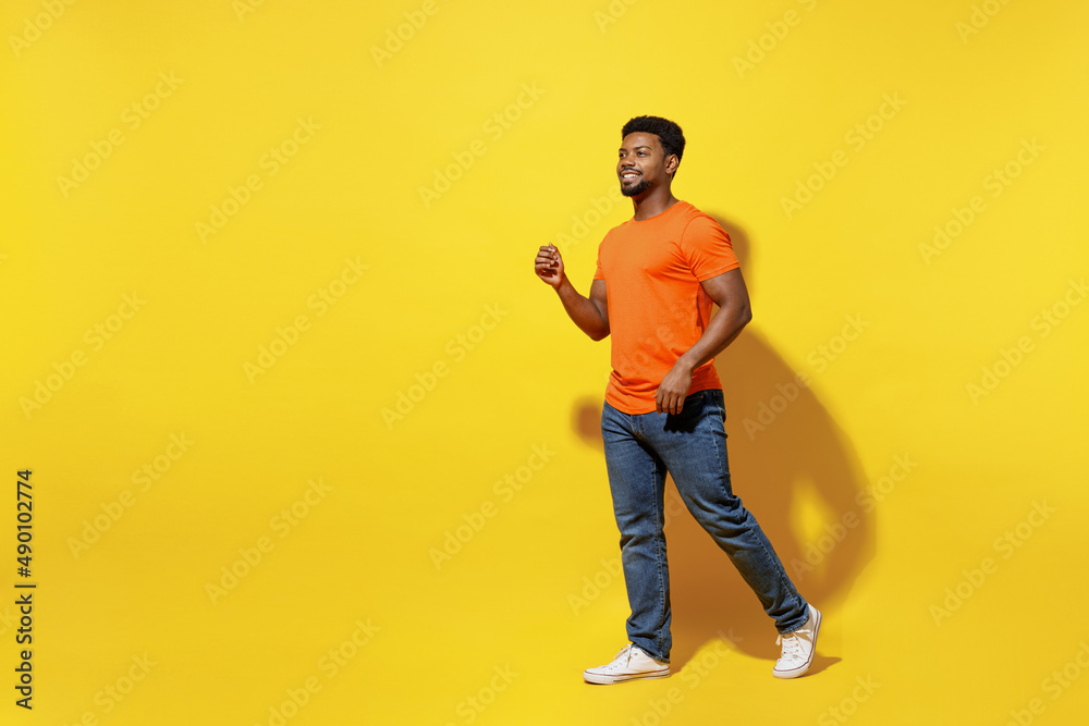 Full body side view smiling happy young man of African American ethnicity 20s wear orange t-shirt walking going strolling isolated on plain yellow background studio portrait. People lifestyle concept. Full body side view smiling happy young man of African American ethnicity 20s wear orange t-shirt walking going strolling isolated on plain yellow background studio portrait. People lifestyle concept.
