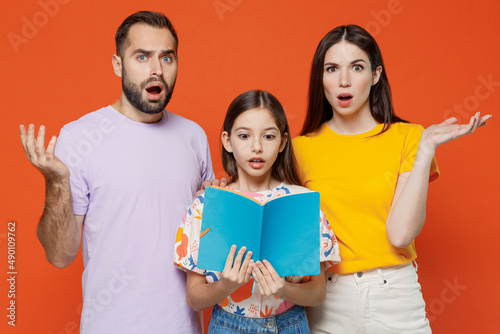 Young confused sad parents mom dad with child daughter teen girl wear basic t-shirts read book with kid help to do hometask spread hands isolated on yellow background. Family day parenthood concept photo