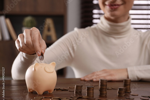 Woman putting coin into piggy bank at wooden table indoors, closeup