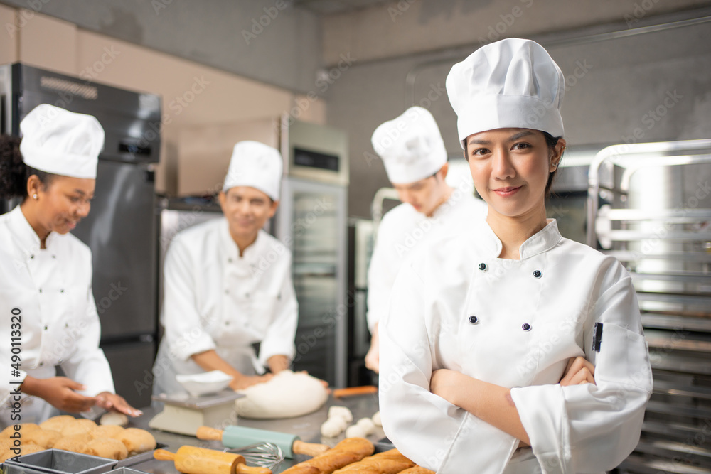 Selective focus of Asian female baker in white chef dress and hat, standing with arms folded and smiling at camera, with blurred colleagues kneading dough in the background. Copy space on left side.