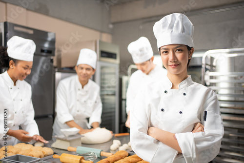 Selective focus of Asian female baker in white chef dress and hat, standing with arms folded and smiling at camera, with blurred colleagues kneading dough in the background. Copy space on left side.