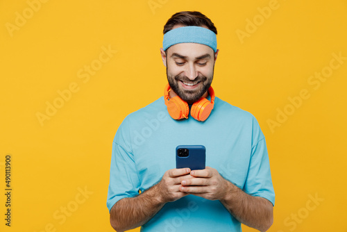 Young fitness trainer instructor sporty man sportsman in headband blue t-shirt spend weekend in home gym hold in hand use mobile cell phone isolated on plain yellow background. Workout sport concept.
