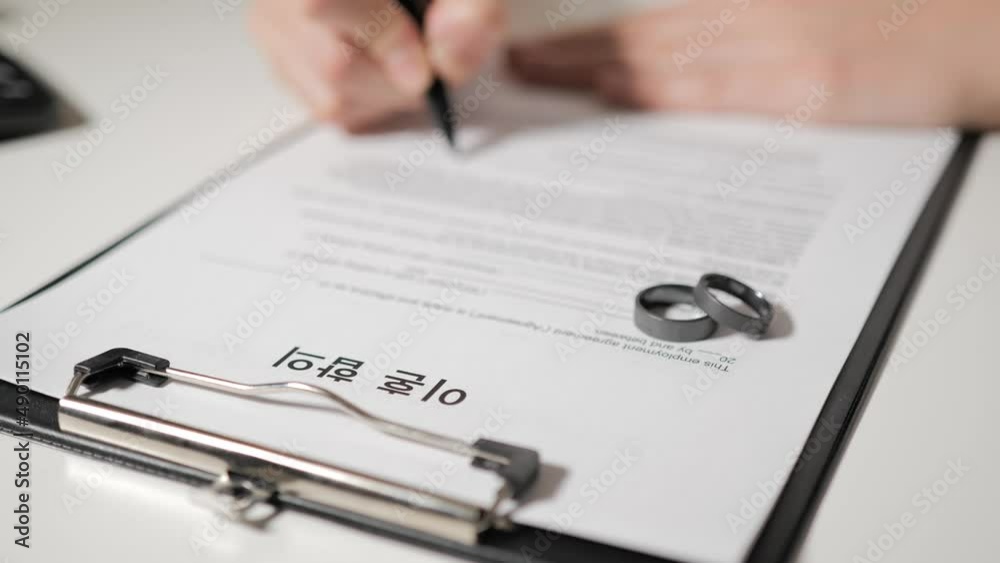 Divorce agreement in Korean language. Hands of husband or wife sign divorce papers. In the foreground are titanium wedding rings. Divorce agreement in Korean language. Hands of husband or wife sign divorce papers. In the foreground are titanium wedding rings.