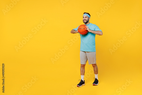 Full body fun young fitness trainer instructor sporty man sportsman in headband blue t-shirt spend leisure time in gym throw basketball ball isolated on plain yellow background. Workout sport concept.